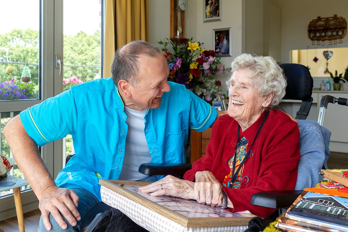 B&BW_medewerker en cliënt_cliënt in rood vest bloemen en lachend in woonkamer