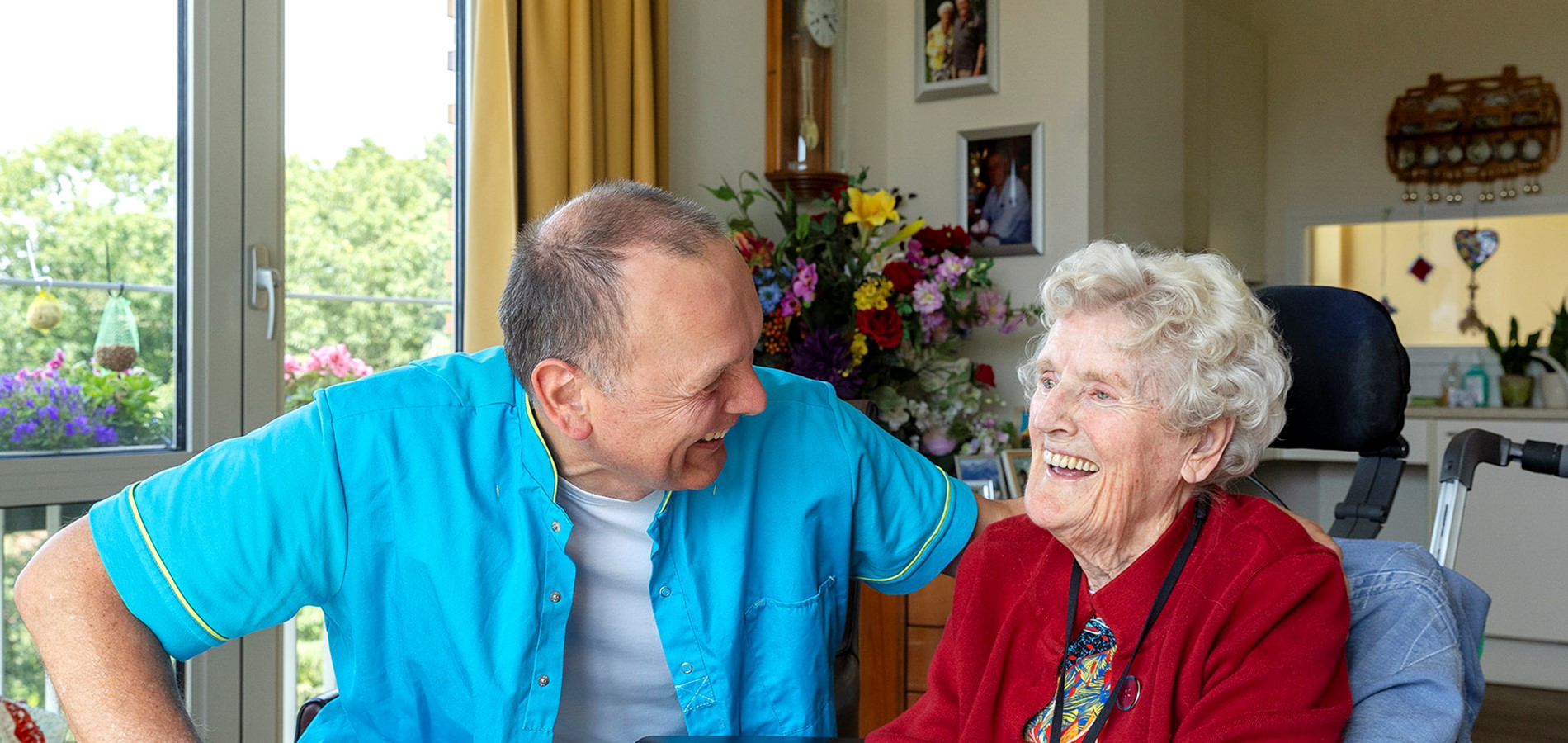 B&BW_medewerker en cliënt_cliënt in rood vest bloemen en lachend in woonkamer