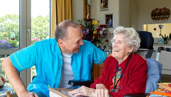 B&BW_medewerker en cliënt_cliënt in rood vest bloemen en lachend in woonkamer