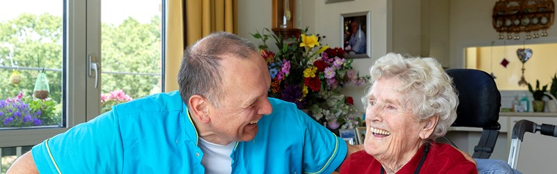 B&BW_medewerker en cliënt_cliënt in rood vest bloemen en lachend in woonkamer
