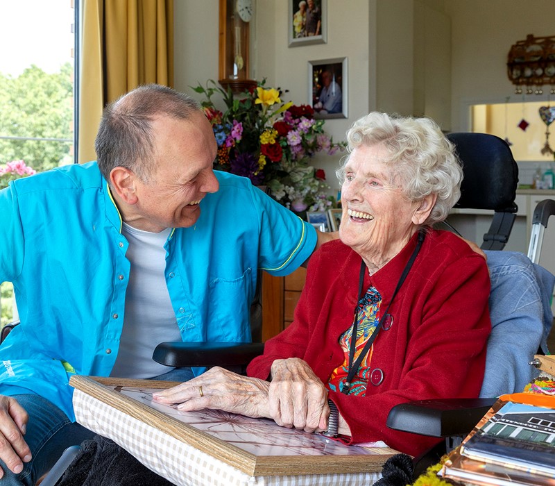 B&BW_medewerker en cliënt_cliënt in rood vest bloemen en lachend in woonkamer