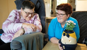 Wijkverpleging_medewerker en cliënt_lachend in stoel in woonkamer met Robot Tessa
